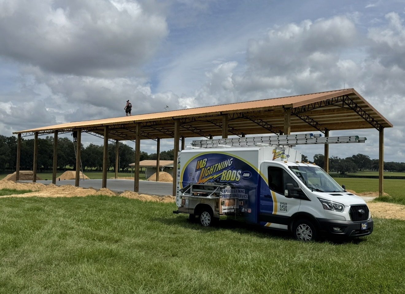 WB Lightning Rods box truck at a large covered riding arena under construction while a crew member works on the roof